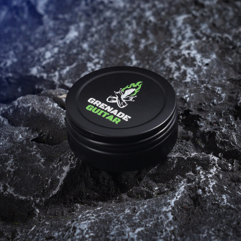 Black Grenade Guitar tin containing assorted Shrapnel V1 picks with neon-green logo on the lid, displayed on a dark textured surface.